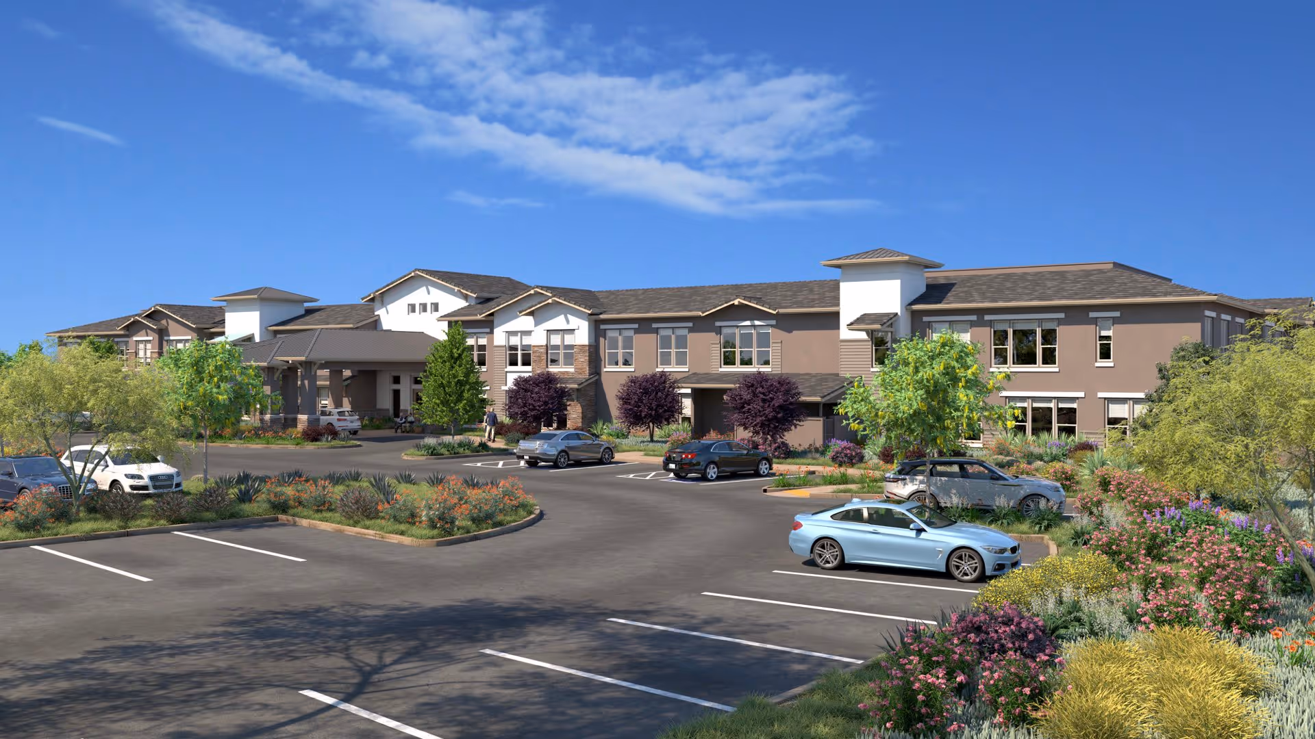 Front exterior of a two-story senior living building with landscaped grounds, a parking lot, and several parked cars under a blue sky.