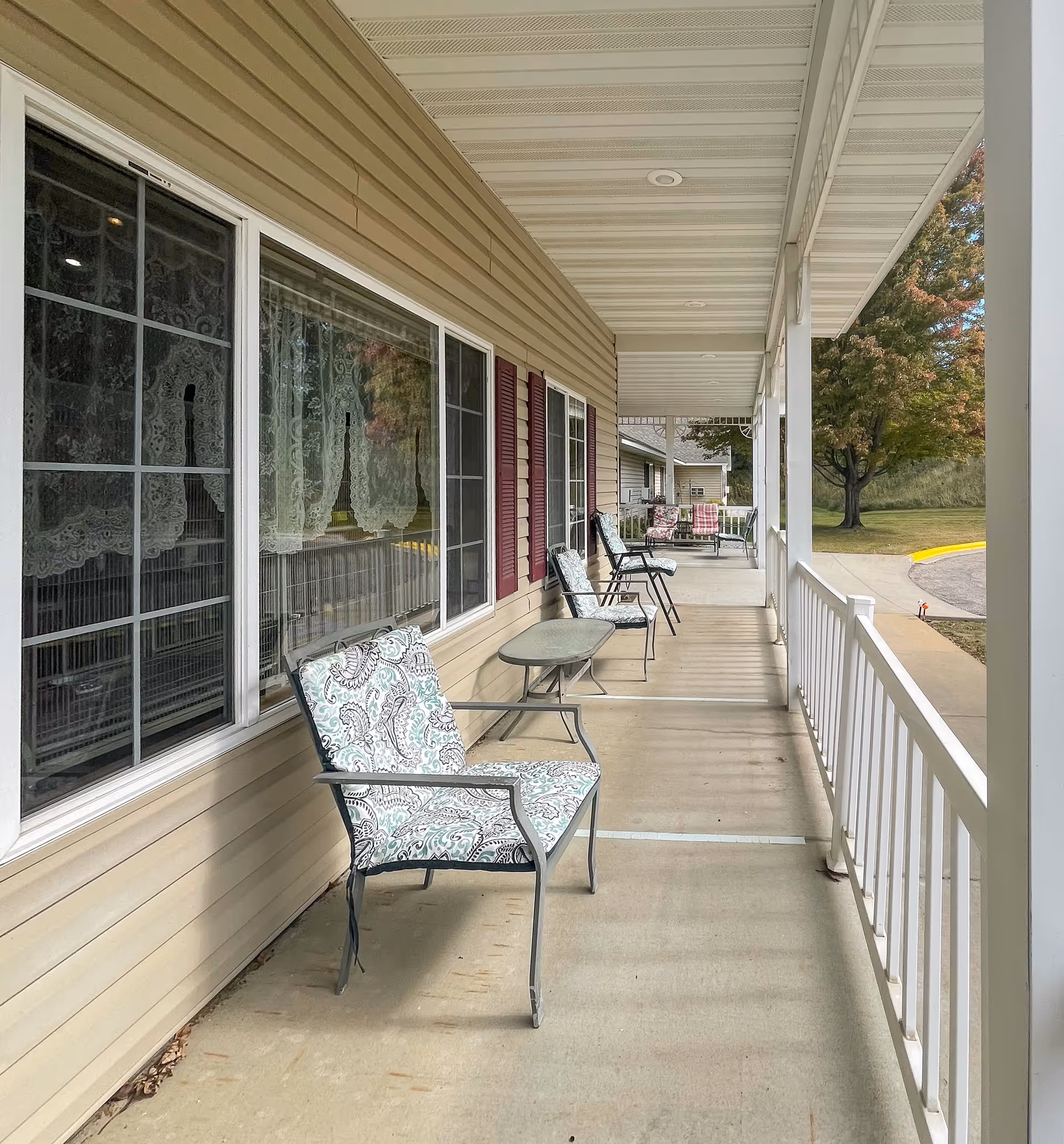 Covered outdoor porch area at Park Lane Estates with several cushioned metal chairs and small tables lined up along the beige siding wall with windows. The porch has a white railing and ceiling, and a tree with autumn foliage is visible in the background.