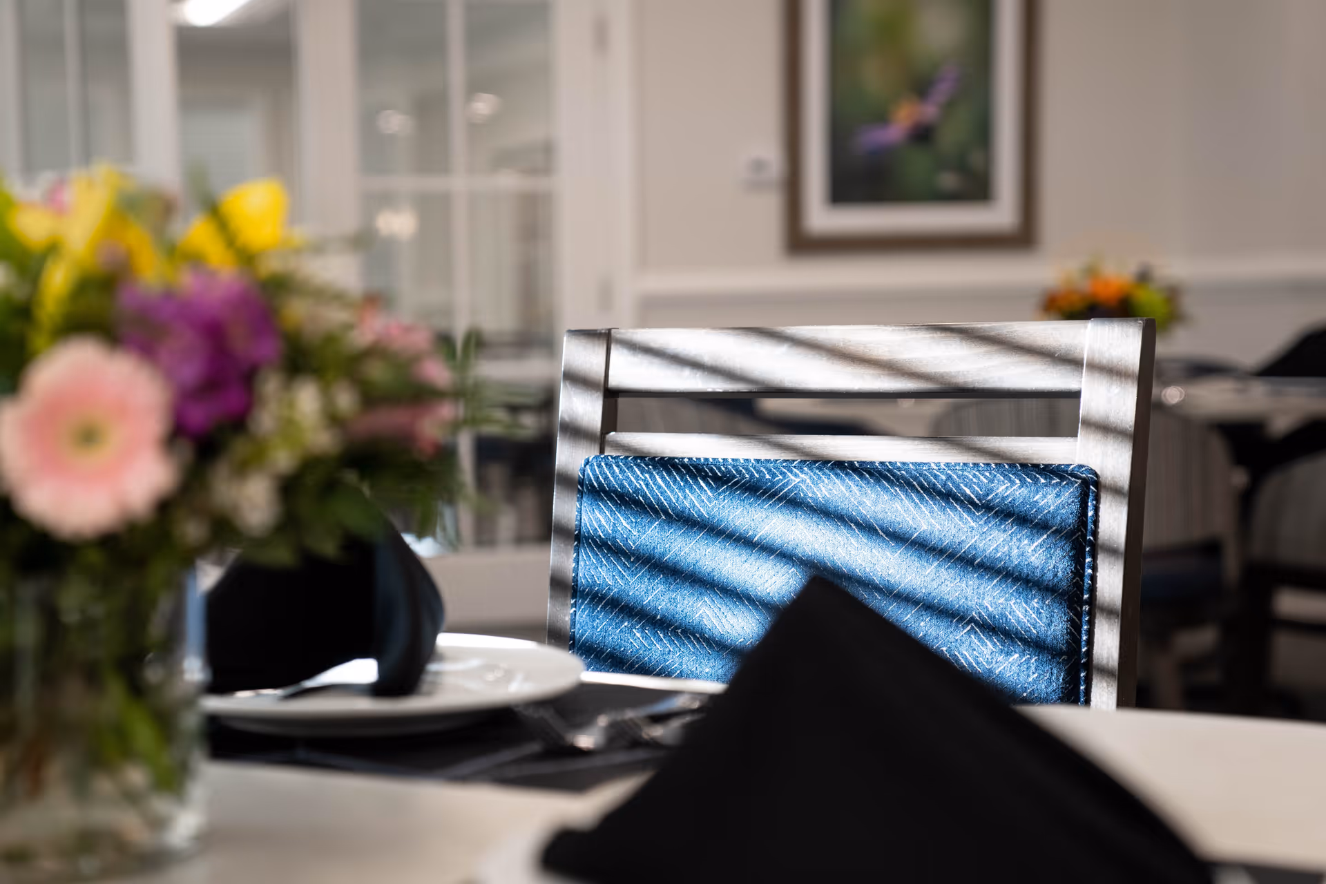 Close-up of a dining chair with a blue cushion in a senior living facility dining room. A table in the foreground is set with a white plate, black napkin, and silverware. A blurred vase with colorful flowers is also visible on the table. The background shows more chairs and a framed picture on the wall.