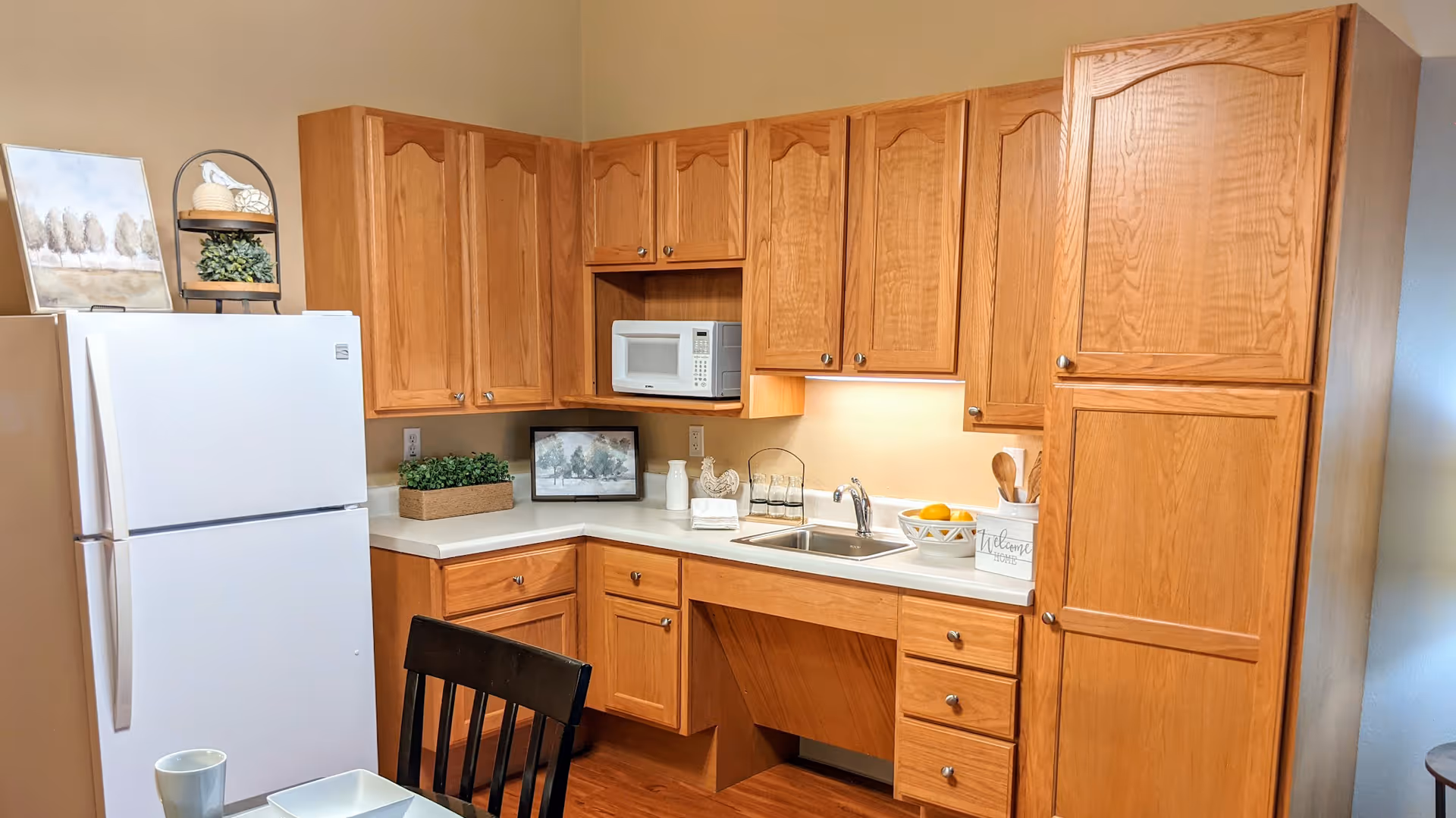 A kitchen area with wooden cabinets, a white refrigerator, a microwave, a sink, and various decorative items including a framed picture, plants, and a bowl of lemons. A black chair and part of a table with a white bowl and a mug are visible in the foreground.
