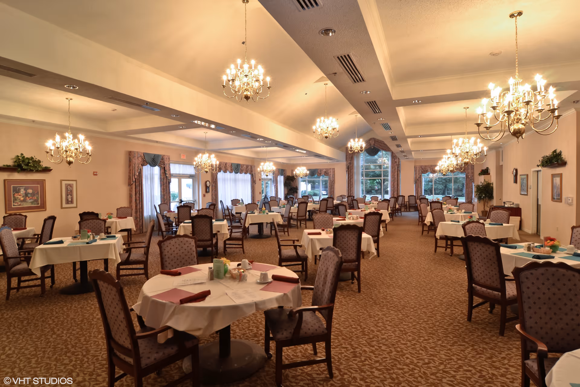 A spacious dining room with multiple round and rectangular tables covered with white tablecloths, each set with napkins, cups, and small centerpieces. The room features elegant chandeliers hanging from the ceiling, patterned carpet, large windows with drapes, and framed artwork on the walls.