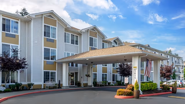 Exterior view of a multi-story senior living facility with a covered entrance driveway, landscaped plants, and an American flag displayed near the entrance.