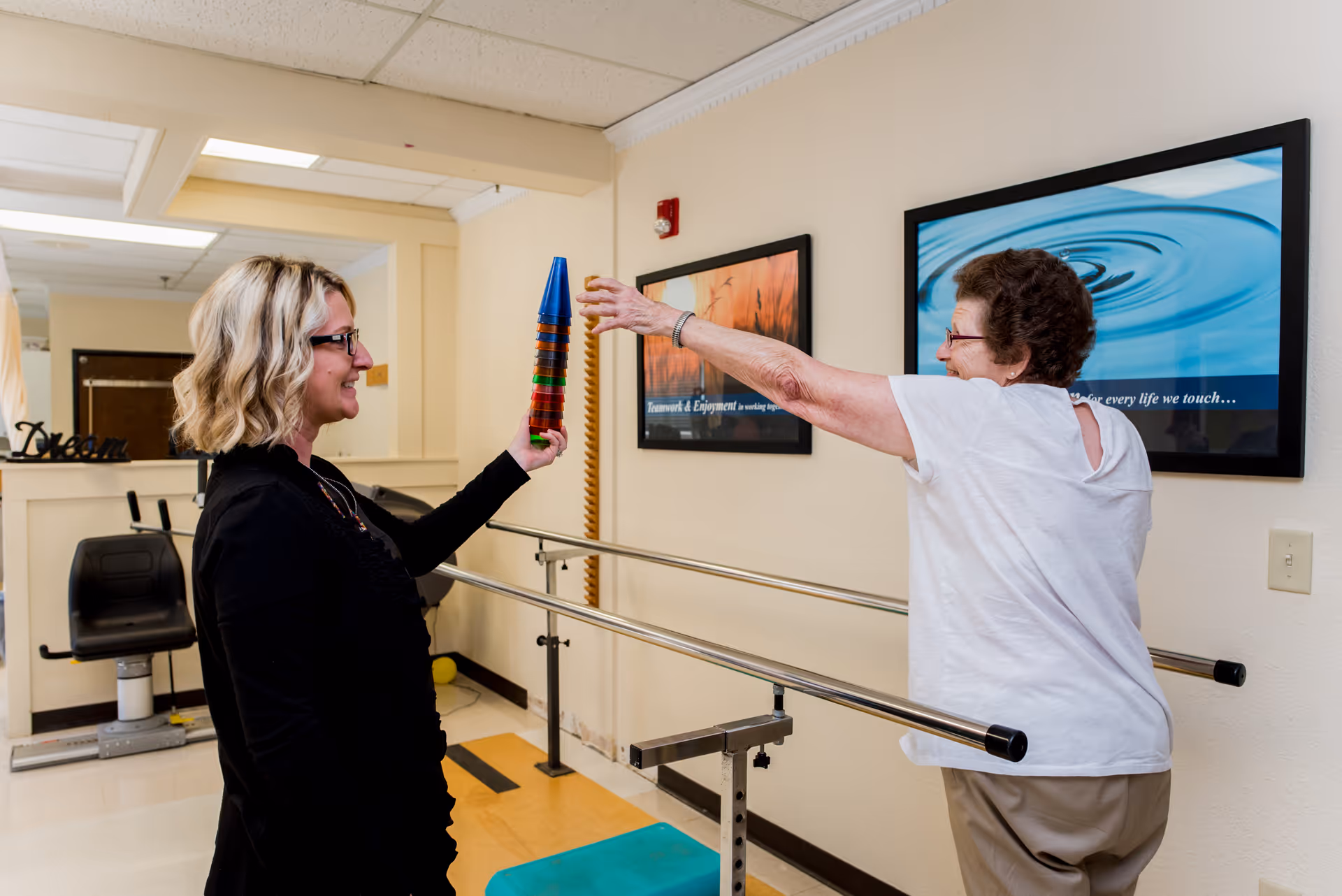 A healthcare worker assists an elderly woman in a physical therapy room. The elderly woman is reaching out to touch a stack of colorful cups held by the healthcare worker. The room has parallel bars for walking support and motivational framed pictures on the wall.