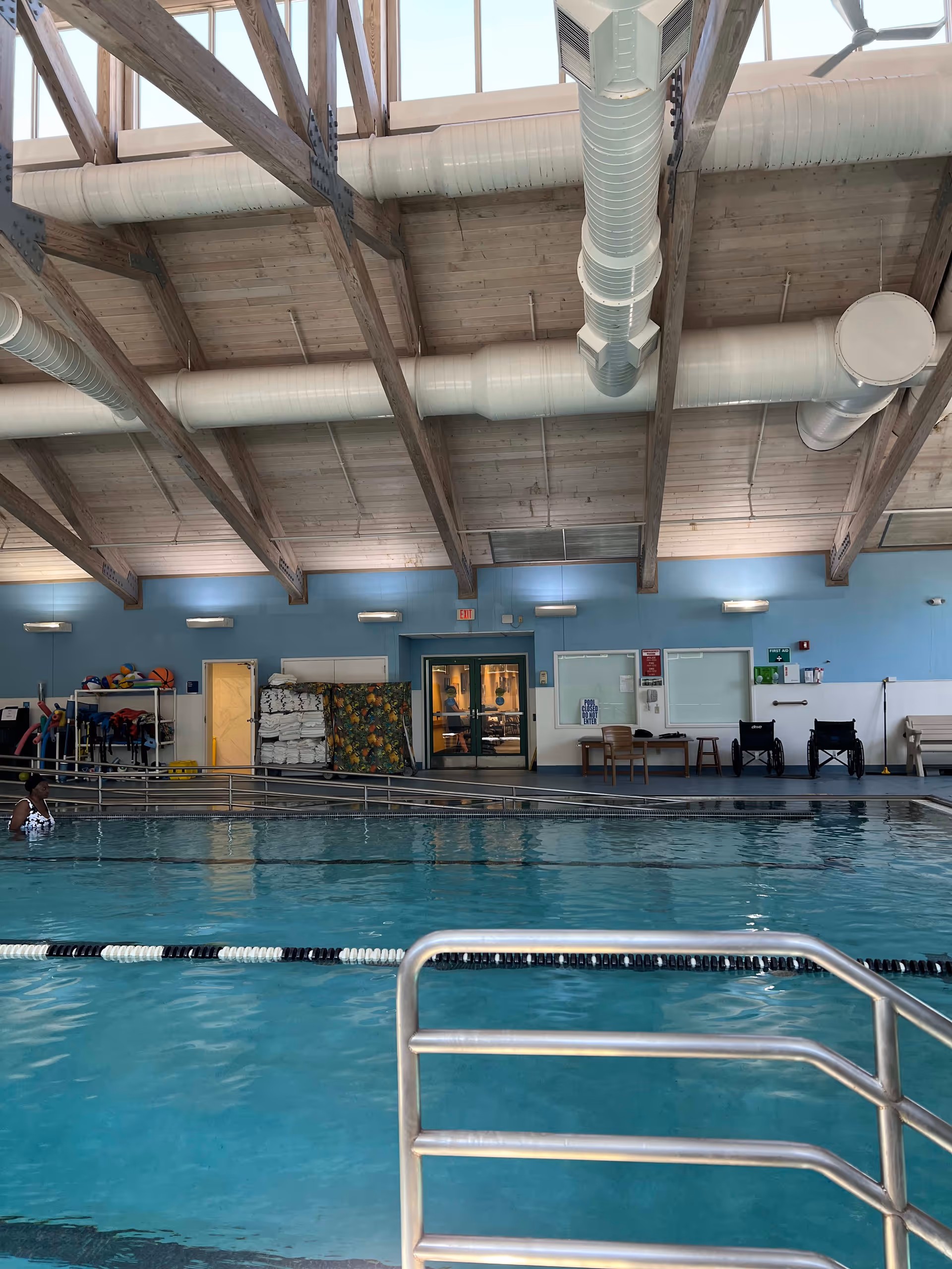 Indoor swimming pool with clear blue water, metal handrails, and a person partially visible in the water on the left side. The pool area has a high ceiling with exposed wooden beams and large white ventilation ducts. The far wall is painted light blue and features double doors, a rack with pool equipment and towels, two wheelchairs, and some chairs and tables.