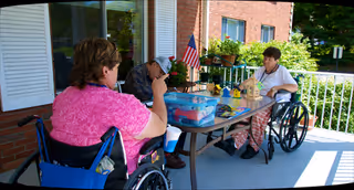 Residents in wheelchairs socialize at a table on a sunny outdoor porch.