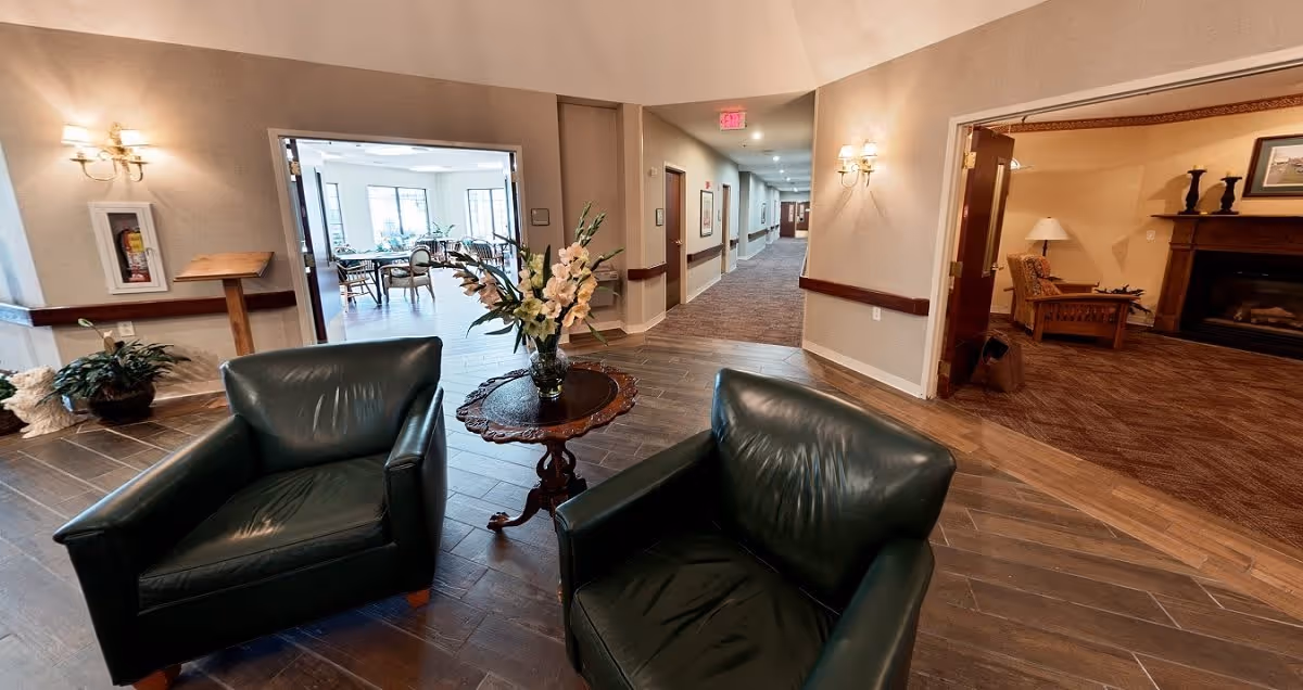 A seating area with two dark green leather armchairs and a round wooden table with a floral arrangement in the center. The area opens into a hallway with carpeted flooring and a dining room with tables and chairs visible through an open doorway. Another room with a fireplace, wooden furniture, and a lamp is visible to the right. The walls are beige with wall sconces providing lighting.
