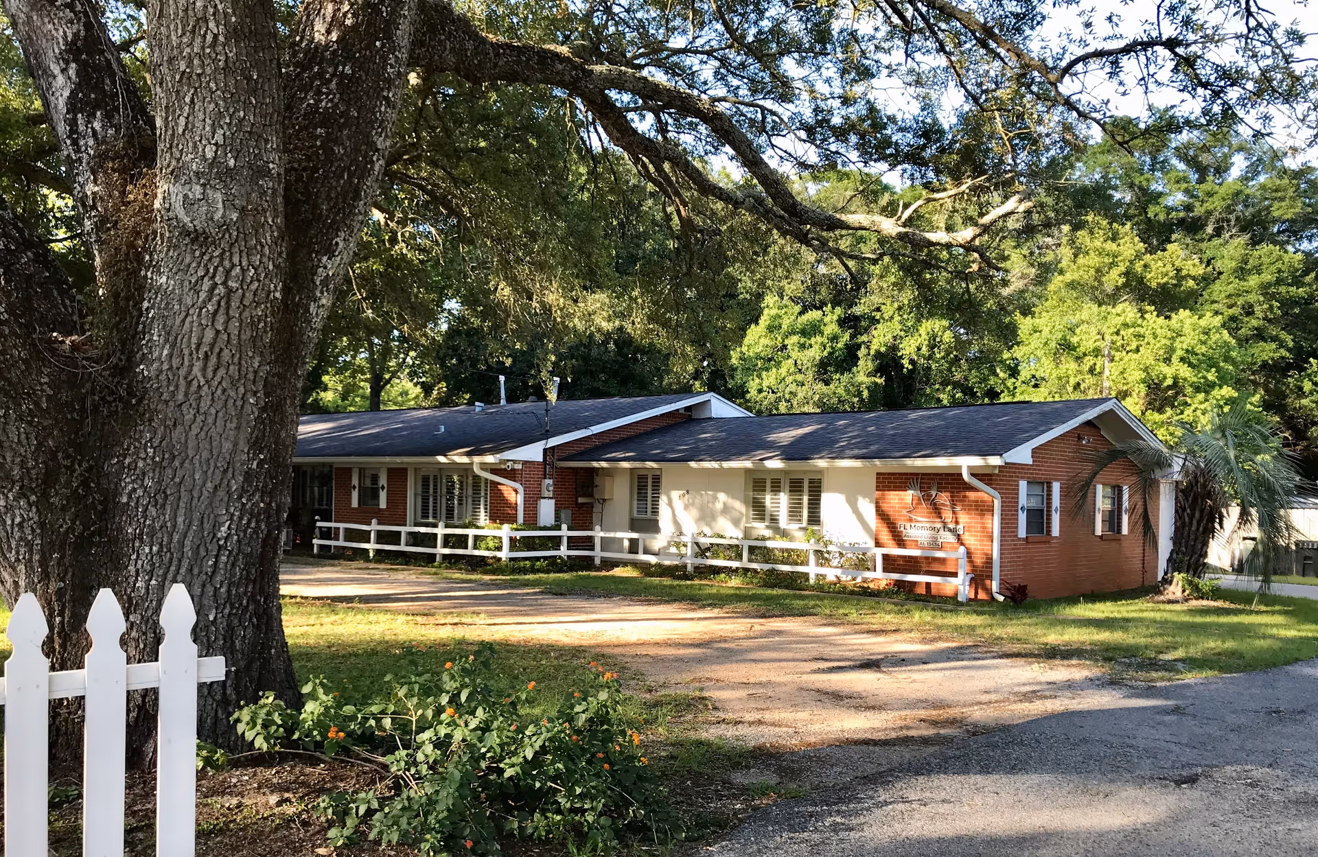 A single-story brick building with white trim and a dark roof, surrounded by trees and greenery. There is a white picket fence and a large tree in the foreground. The building has a sign that reads 'FL Memory Lane' and appears to be a residential or care facility.