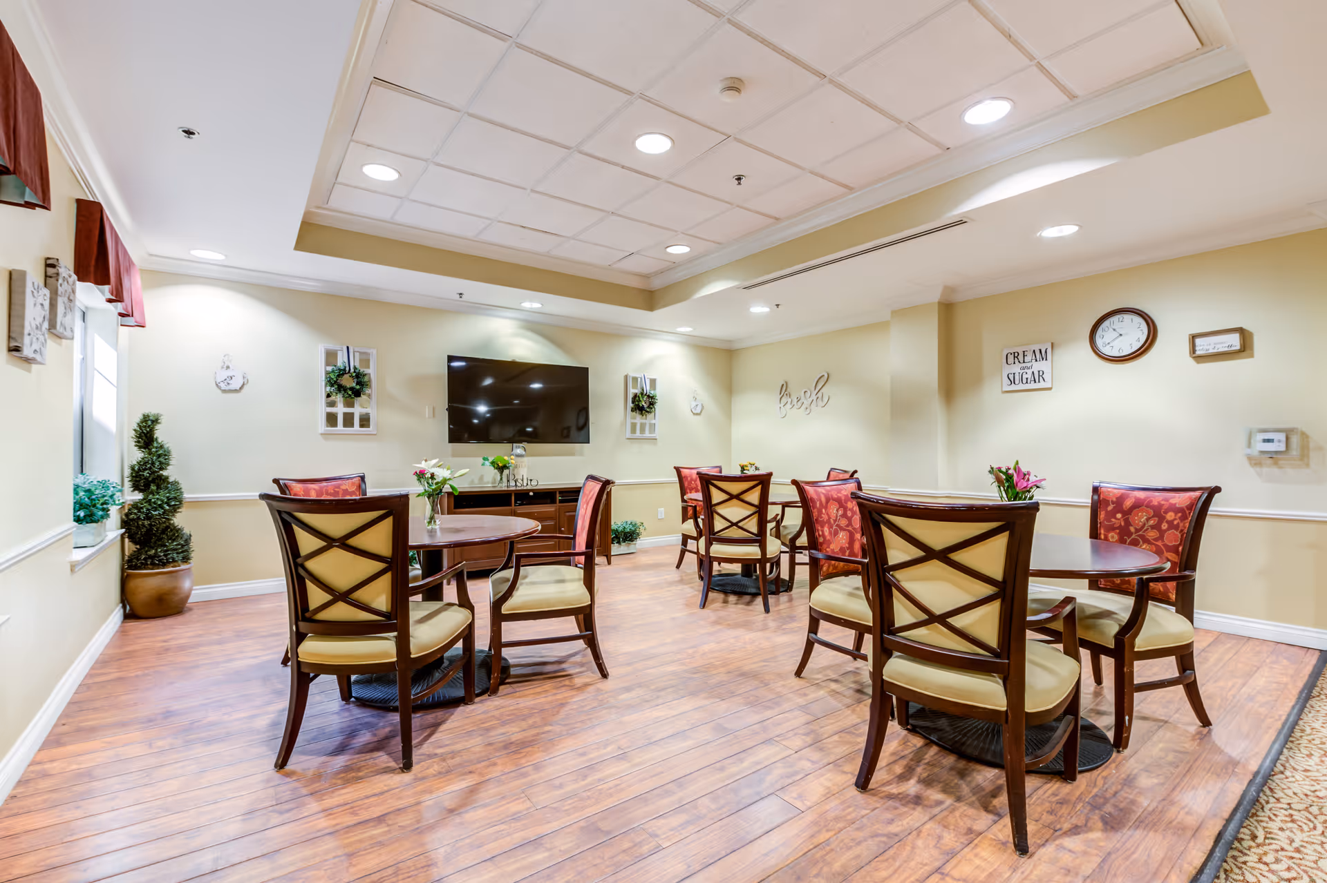 A bright and clean common area in a senior living facility with several round wooden tables and cushioned chairs arranged around the room. The walls are light yellow with decorative wall art, a clock, and a flat-screen TV mounted on one wall. There are small plants and flowers on the tables and windowsills, and the floor is a wood-like laminate.