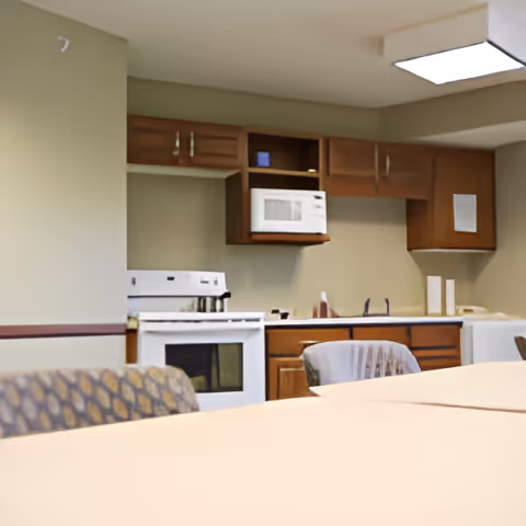 Interior view of a kitchen area in a senior living facility with wooden cabinets, a white microwave, a white stove, and a sink. In the foreground, there are tables and chairs with patterned upholstery.