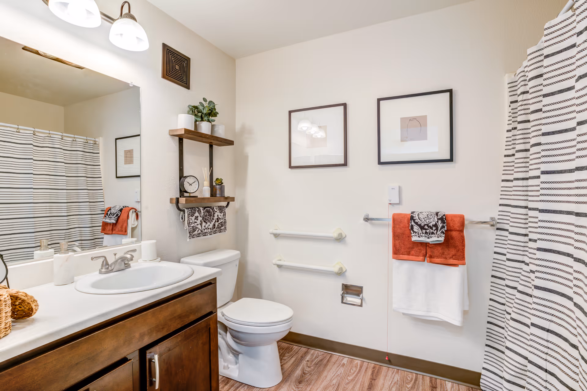 A clean and well-lit bathroom featuring a white sink with a silver faucet set in a wooden vanity. Above the sink is a large mirror with two light fixtures. To the right is a white toilet with two wooden shelves above it holding small plants and decorative items. On the wall are two framed pictures and two grab bars for support. A towel rack holds folded red and white towels with a patterned cloth on top. A striped shower curtain is visible on the right side of the image. The floor has a wood-like finish.