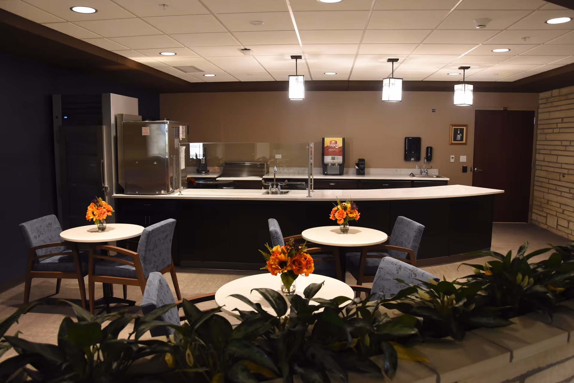 A small dining area with three round tables, each decorated with a vase of orange flowers. The tables are surrounded by gray upholstered chairs with wooden armrests. Behind the tables is a kitchen counter with a sink, a coffee dispenser, and other kitchen appliances. The room has a beige and dark blue color scheme with recessed lighting and pendant lights hanging from the ceiling. There are green plants in the foreground.