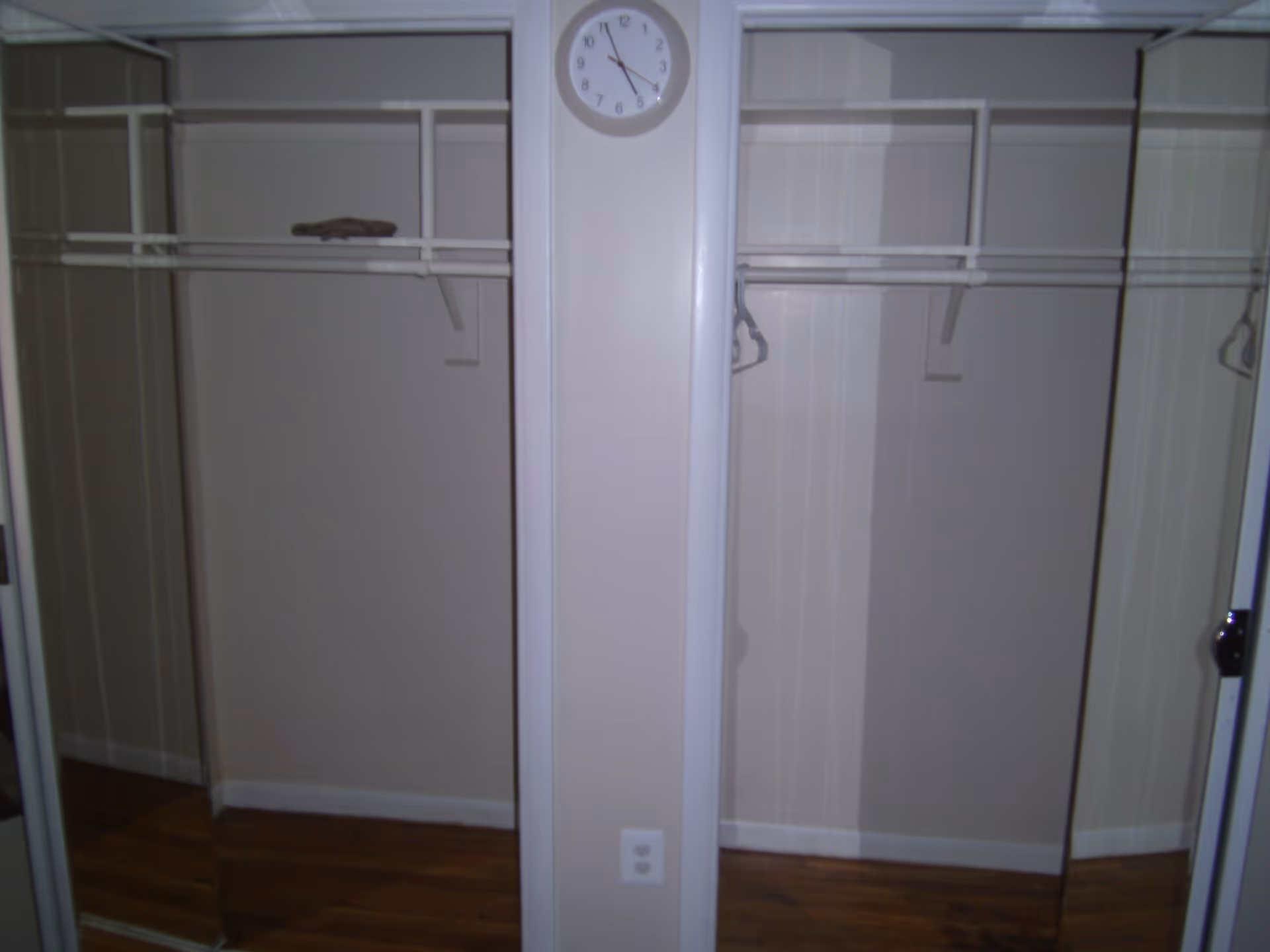 Empty closet with mirrored sliding doors on both sides, a white wall with a clock above an electrical outlet in the center, and wooden flooring.