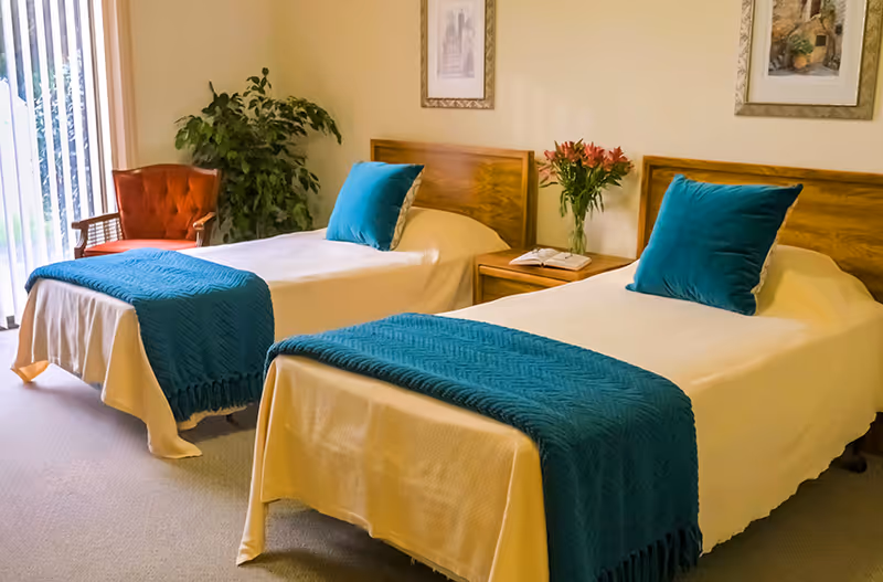 Bright bedroom with two twin beds made with white linens and teal throws, a nightstand with flowers, a chair, and a sliding glass door.