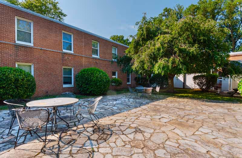 Outdoor patio area with stone flooring, metal tables and chairs, surrounded by greenery including bushes and a large tree, adjacent to a two-story brick building with multiple windows.