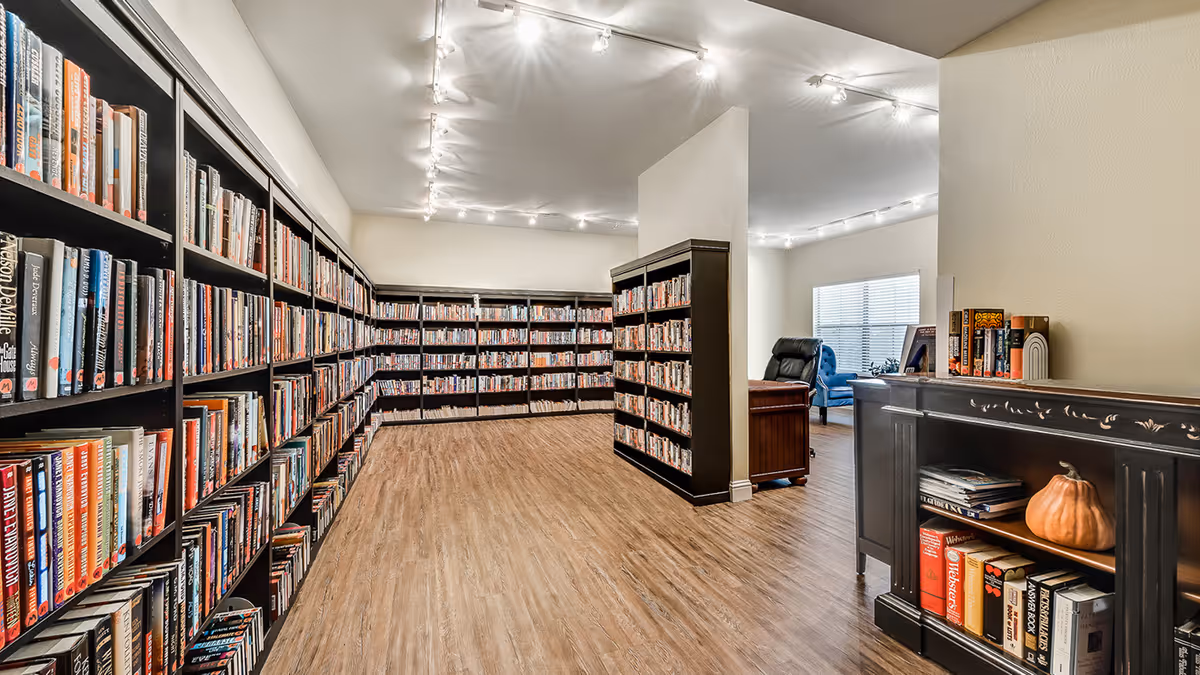 Interior view of a well-lit library room with wooden floors and multiple black bookshelves filled with books. There is a small black cabinet with books and a decorative pumpkin on the right side. In the background, there is a seating area with a black leather chair and a blue armchair near a window with blinds.
