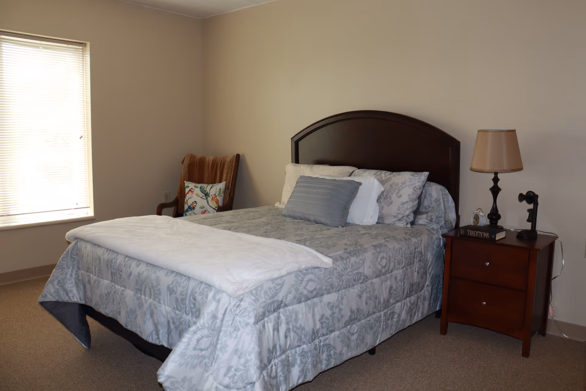 A bedroom with a neatly made bed featuring a gray patterned comforter and multiple pillows. To the right of the bed is a wooden nightstand with a lamp, a book, and a vintage-style telephone. In the corner near the window, there is a wooden chair with a brown throw blanket and a decorative pillow with a bird design. The room has beige walls and carpeted flooring.