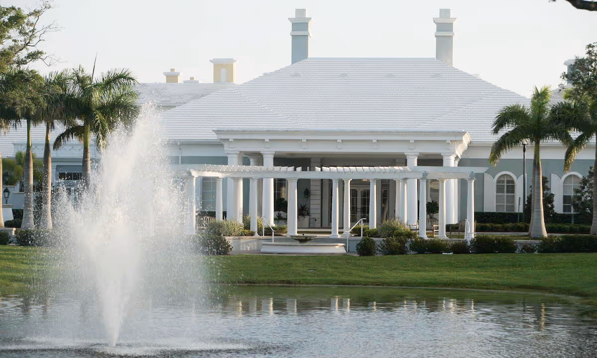 View of a large white building with a white roof and columns, surrounded by palm trees and greenery, with a water fountain spraying in a pond in the foreground.
