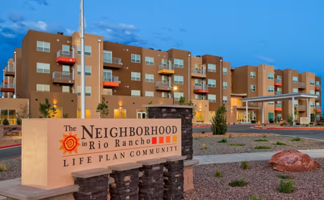 Front exterior of a multi-story senior living community with a prominent sign reading "The Neighborhood in Rio Rancho Life Plan Community."
