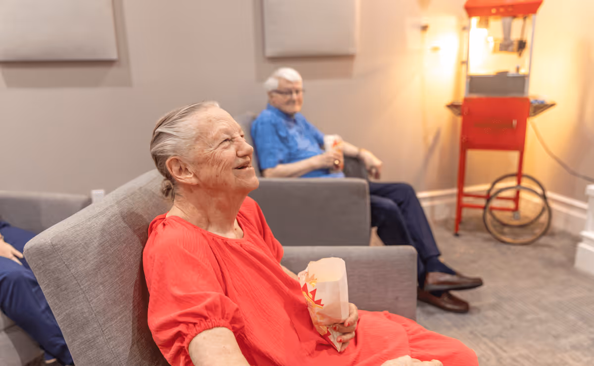 Two elderly individuals sitting in a lounge area, each holding a bag of popcorn. One person in a red shirt is smiling and looking to the side, while the other in a blue shirt is seated further back. A red popcorn machine is visible in the background.