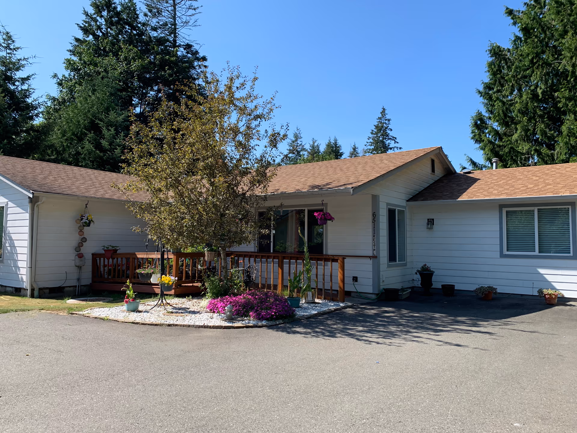 Exterior view of a single-story white building with a brown roof, surrounded by trees and a clear blue sky. There is a small garden area with flowers and a tree in front of the building, along with a wooden railing and a paved driveway.
