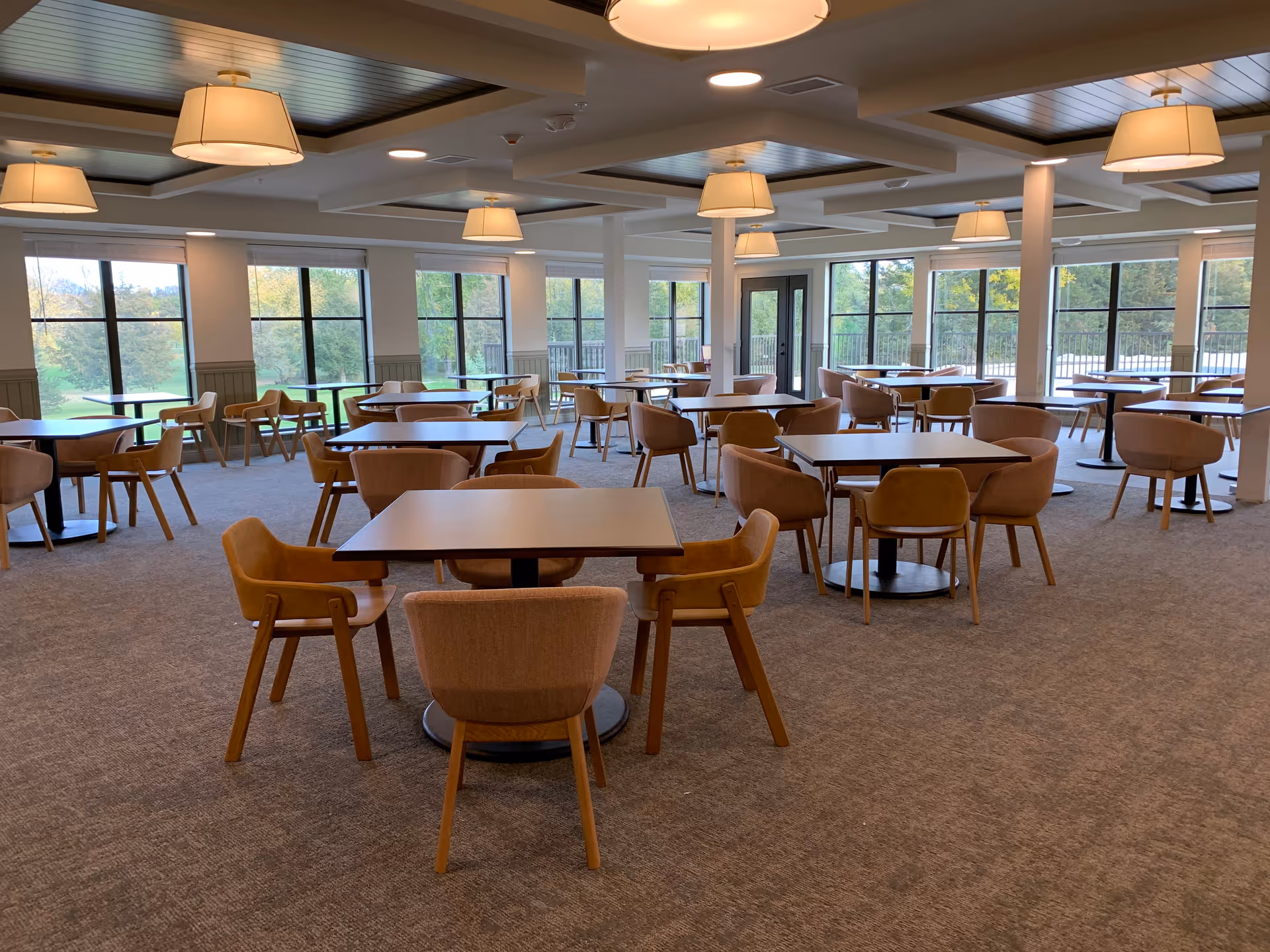 A spacious dining room with multiple square tables and cushioned chairs arranged neatly. Large windows surround the room, allowing natural light to fill the space and offering views of greenery outside. The ceiling features recessed lighting and hanging lamps.