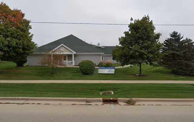 Single-story light-gray building with a green roof set back behind a grassy lawn and trees and a small sign near the sidewalk.
