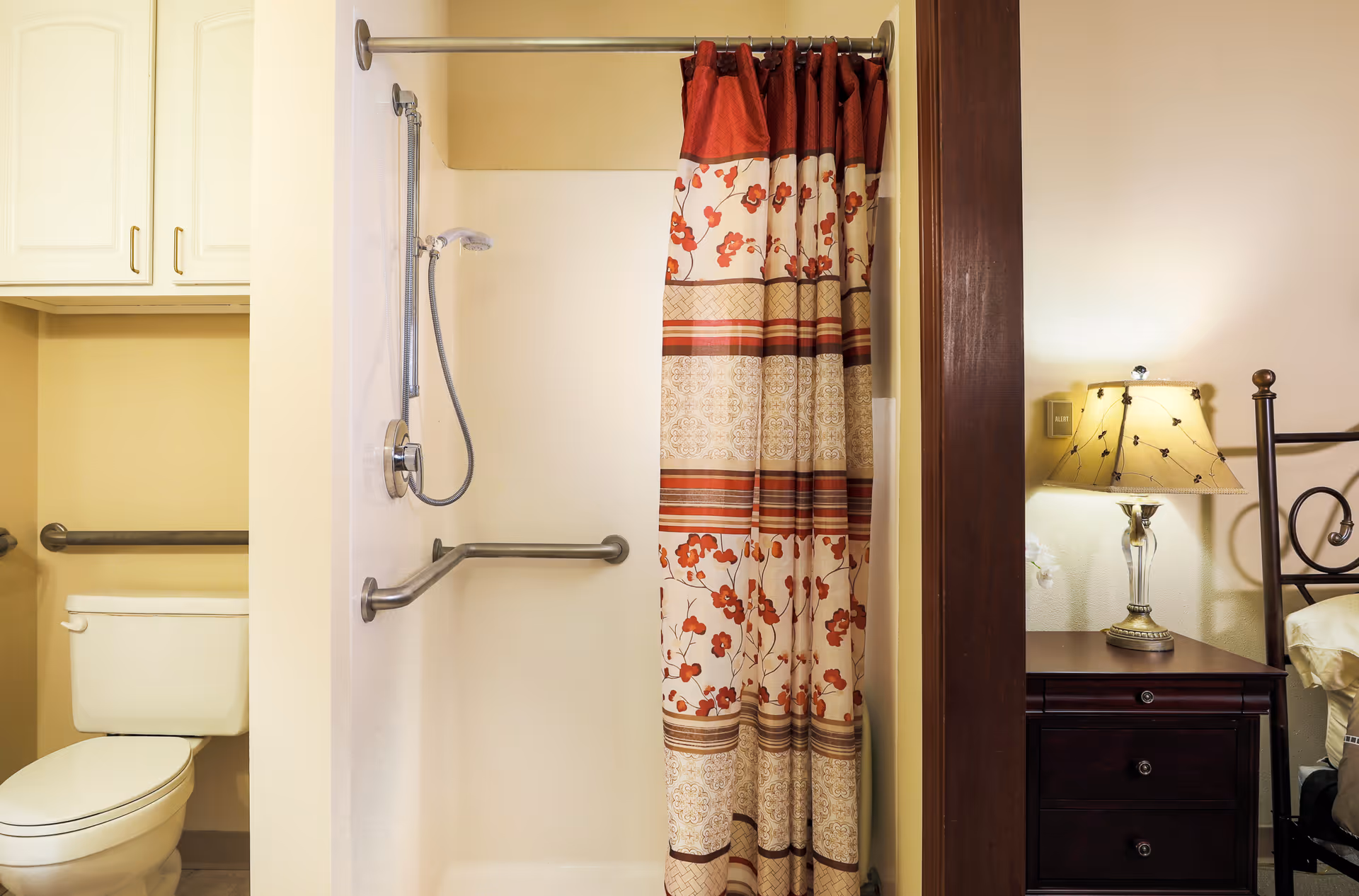 A bathroom with a white toilet and grab bars, a shower with a floral and striped shower curtain, and a glimpse of a bedroom with a nightstand and lamp visible through an open doorway.