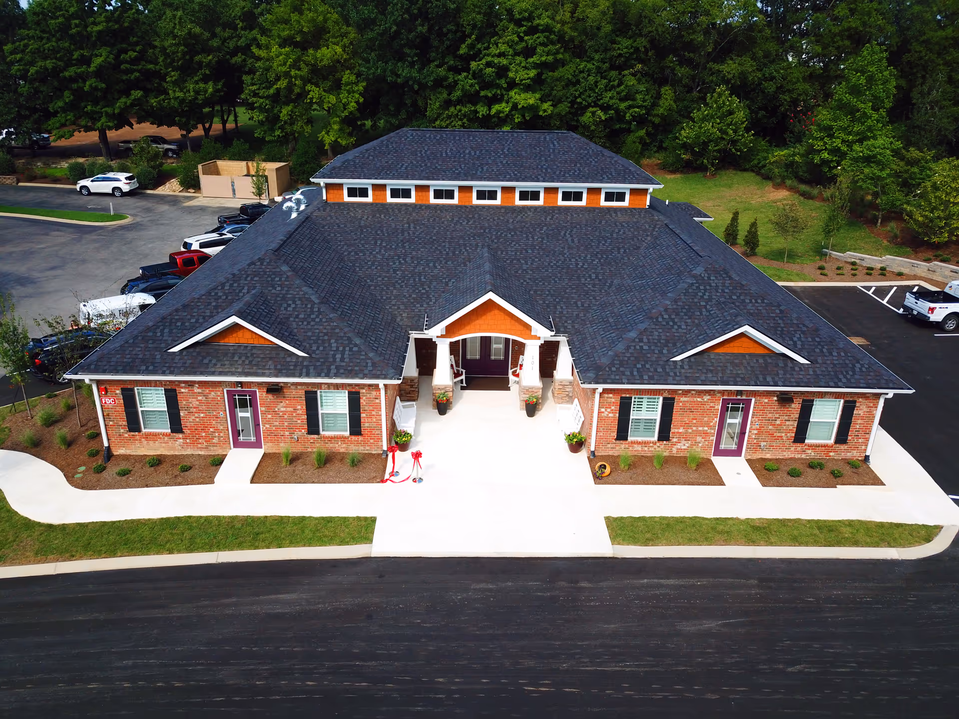 Aerial front view of a single-story brick memory care building with a dark roof, central entrance, parking and surrounding landscaping.