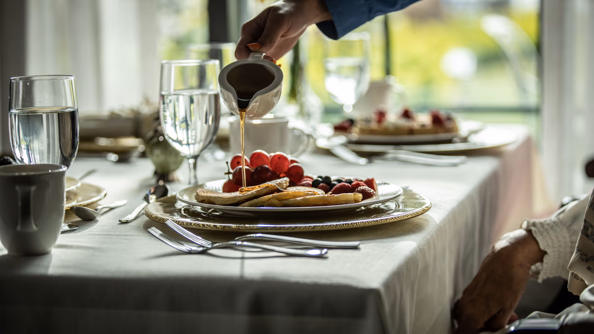 A hand pours syrup onto pancakes and fruit on a plated breakfast at a set dining table.