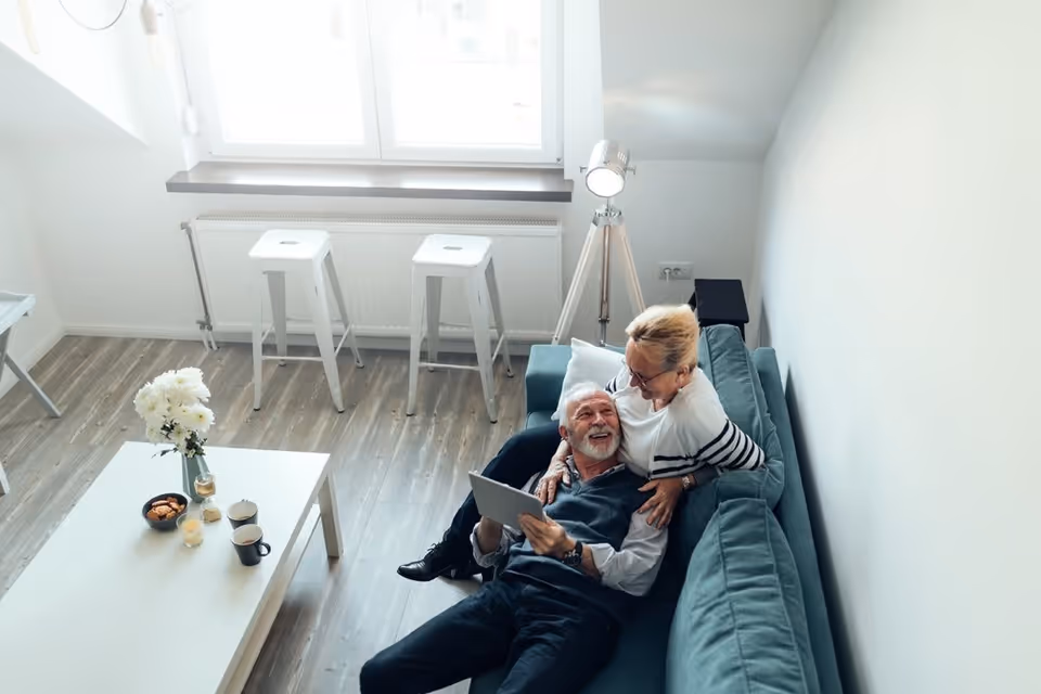 An elderly couple sitting on a teal sofa in a bright living room. The man is holding a tablet and smiling while the woman leans over him affectionately. The room has wooden flooring, a white coffee table with flowers, snacks, and drinks, two white stools near a window, and a tripod floor lamp.