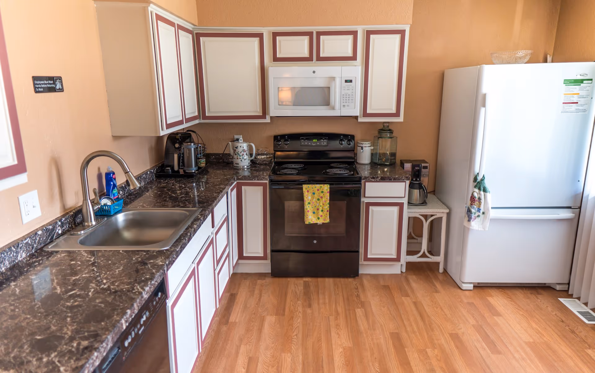 A kitchen with white cabinets trimmed in maroon, a black electric stove with a yellow polka dot towel hanging on the handle, a white microwave above the stove, a white refrigerator with a decorative towel hanging on the handle, a stainless steel sink, a coffee maker, and a wooden floor.