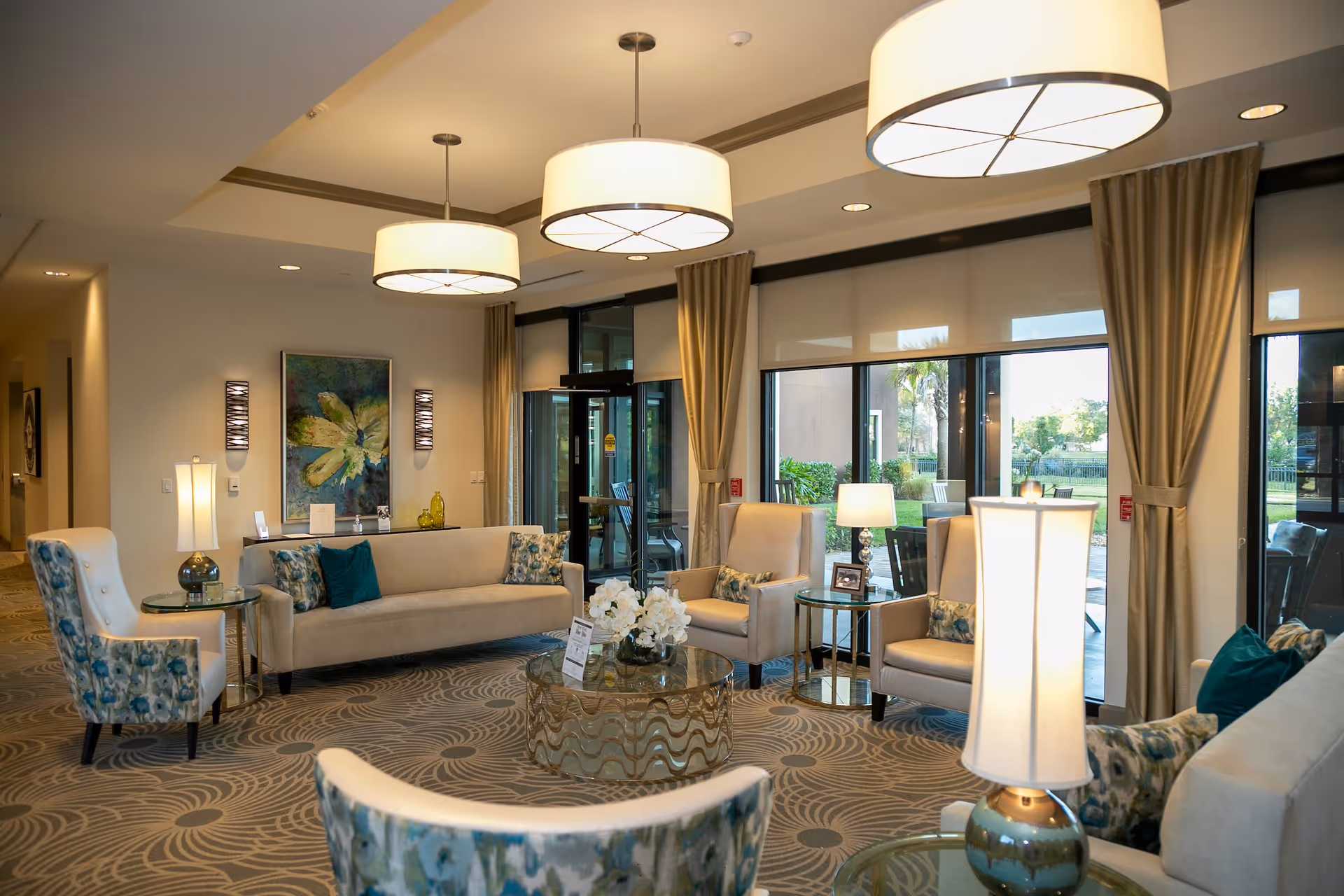 A well-lit living room area in a senior living facility with beige sofas and armchairs featuring blue floral patterns. The room has large windows with beige curtains, round glass coffee tables with decorative items, and modern ceiling lights. There is a painting on the wall and a view of an outdoor patio area through the windows.