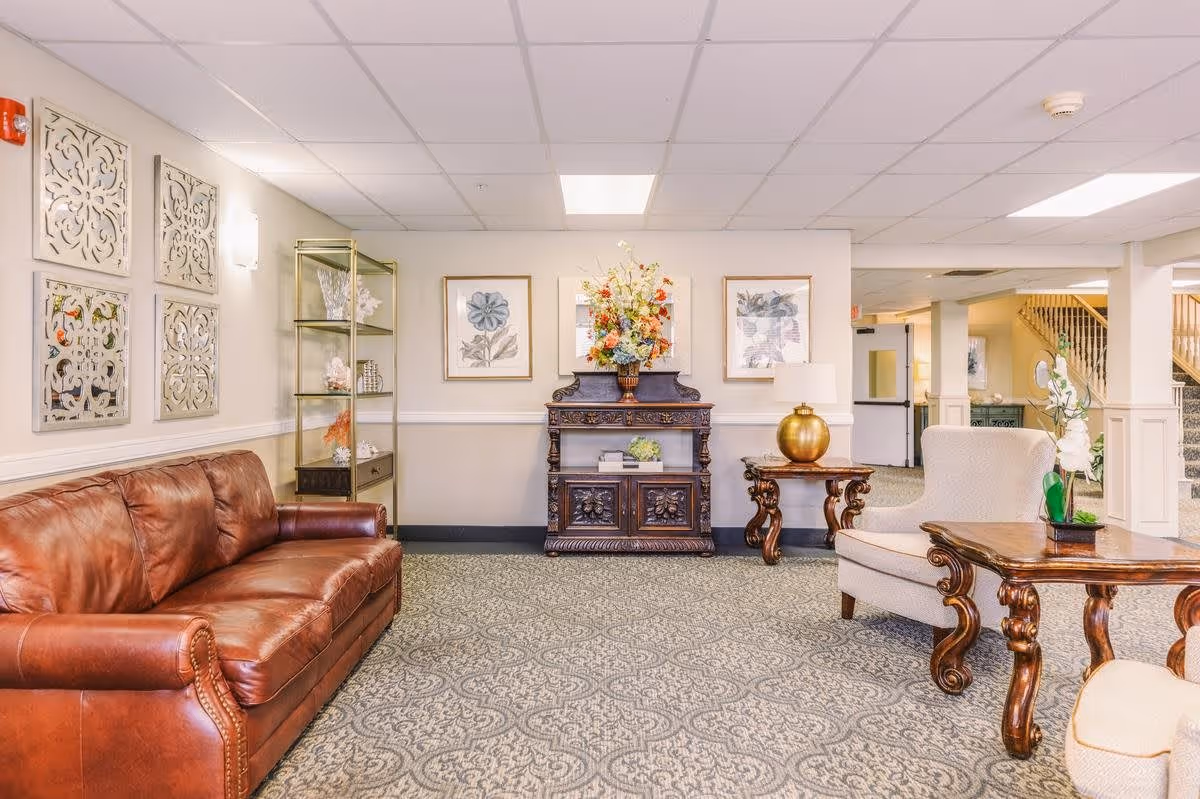 A cozy senior living facility common area with a brown leather sofa on the left, a beige armchair on the right, and ornate wooden tables. The back wall features floral artwork, a decorative wooden cabinet with a large flower arrangement, and a glass display shelf with decorative items. The carpet has a patterned design, and the ceiling has recessed lighting panels.
