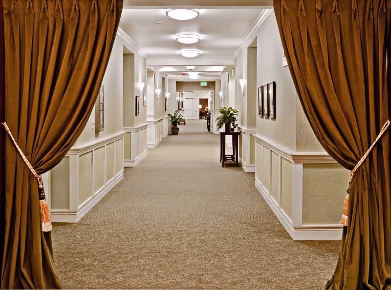 A long, well-lit hallway in a senior living facility with beige carpet and cream-colored walls. The hallway is framed by two large brown curtains tied back with tassels. There are several ceiling lights, wall sconces, framed pictures on the walls, and potted plants placed along the corridor.