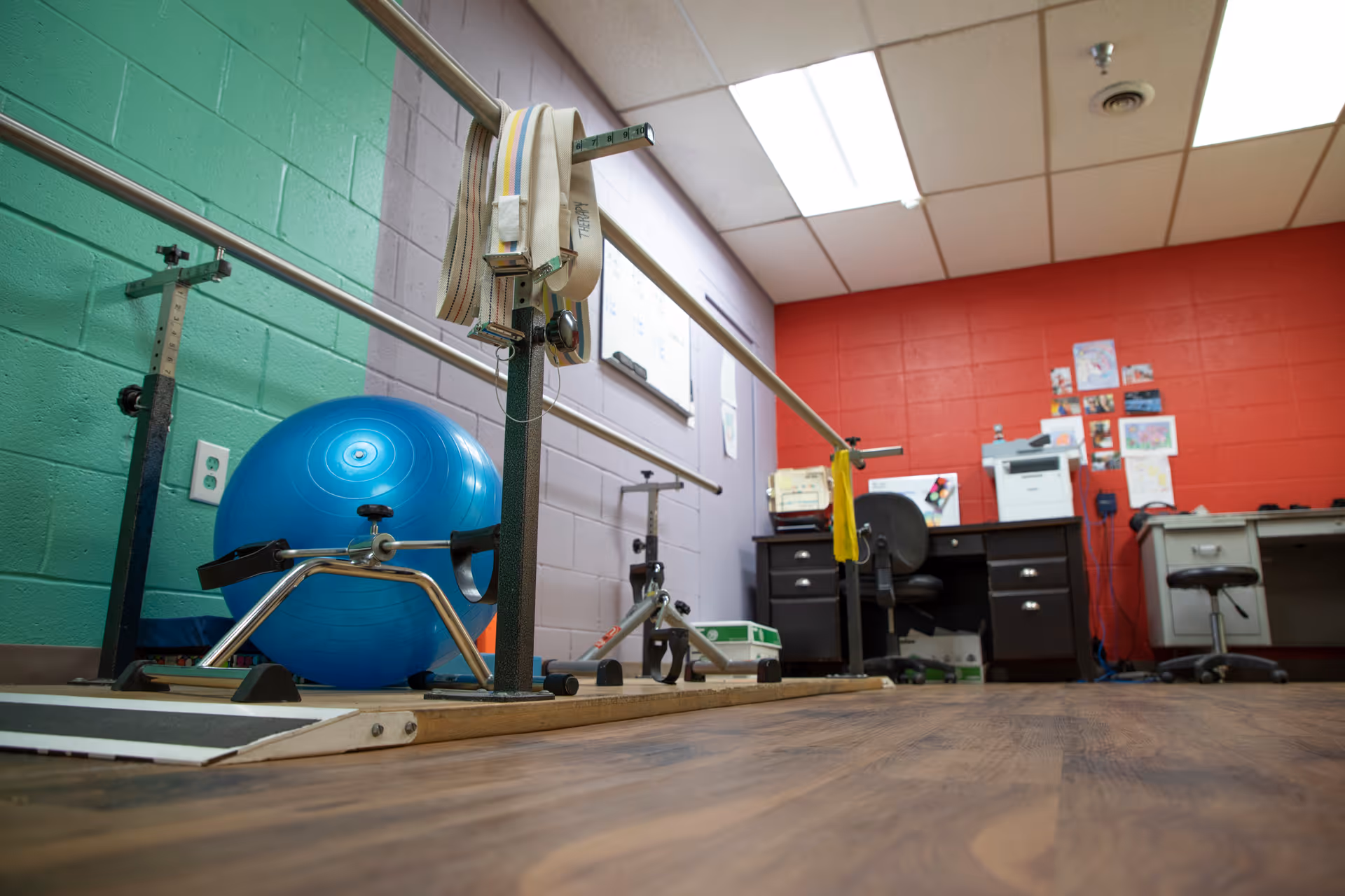 Low-angle view of a rehab therapy room with parallel bars, a blue exercise ball, desks and office equipment against colorful painted cinderblock walls.