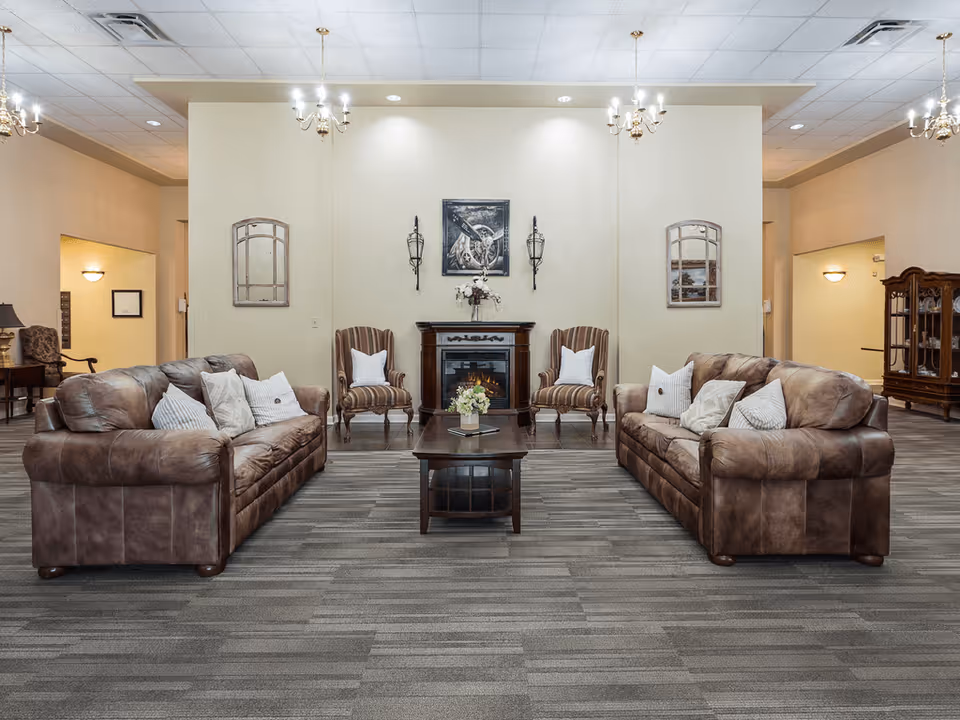 A cozy living room area in a senior living facility featuring two brown leather sofas facing each other with white and striped pillows. Between them is a dark wooden coffee table with a small flower arrangement and a book. Behind the coffee table are two striped armchairs with white pillows, flanking a decorative electric fireplace with a floral arrangement on top. The walls are light-colored with two wall sconces and framed artwork above the fireplace. The room has a gray carpeted floor and is softly lit by chandeliers and wall lights.