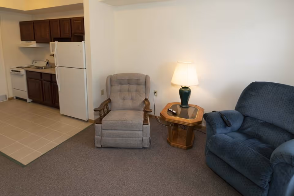 A small living area with a gray upholstered armchair and a blue recliner chair separated by a wooden side table with a green lamp on it. In the background, there is a kitchenette with dark wooden cabinets, a white refrigerator, a stove, and a sink. The floor is carpeted in the living area and tiled in the kitchen area.