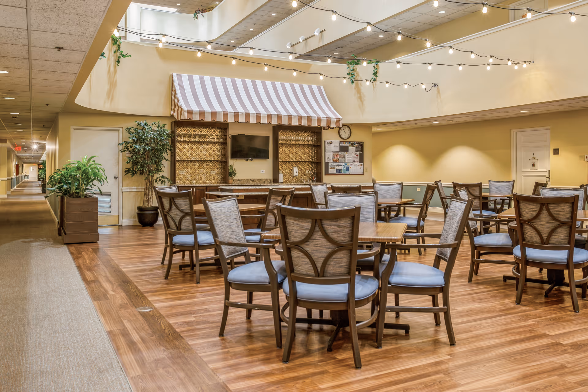 A spacious indoor dining area with multiple wooden tables and chairs with blue cushions. The room has warm yellow walls, wooden flooring, and a striped awning over a counter area with a mounted TV. String lights hang from the ceiling, and there are potted plants near the walls. A hallway extends to the left side of the room.