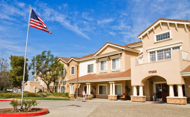 Exterior view of a two-story senior living facility building with beige walls and a tiled roof under a blue sky with some clouds. An American flag is flying on a flagpole in front of the building, which has a covered entrance supported by columns. Trees and landscaping surround the area.