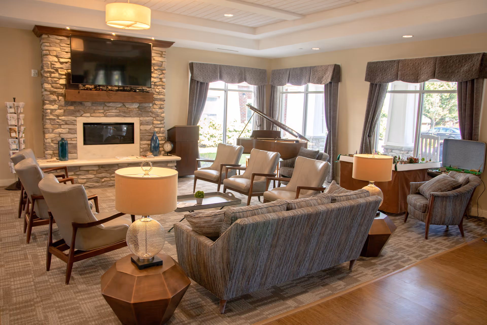 Sunny communal living room with sofas and armchairs arranged around a fireplace and TV, lamps, and a grand piano by large windows.