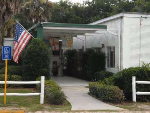Entrance to a white nursing home with green awning, flag, and shrubs.