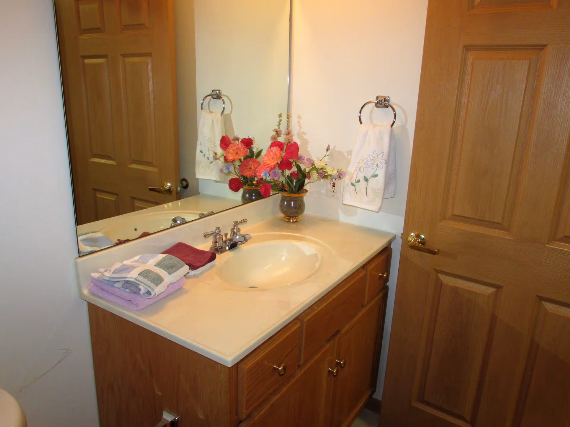 Bathroom vanity with a sink, a large mirror, wooden cabinets, a vase with colorful flowers, folded towels on the counter, and a towel hanging on a ring on the wall next to a wooden door.