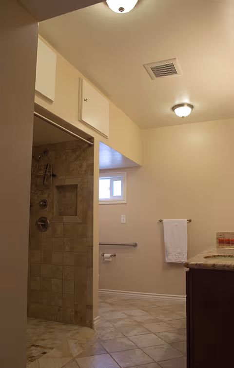 Interior view of a bathroom in a residential care facility featuring a tiled walk-in shower with a built-in niche, a towel hanging on a wall-mounted rack, a small window, a grab bar near the toilet paper holder, and a granite countertop with a sink.