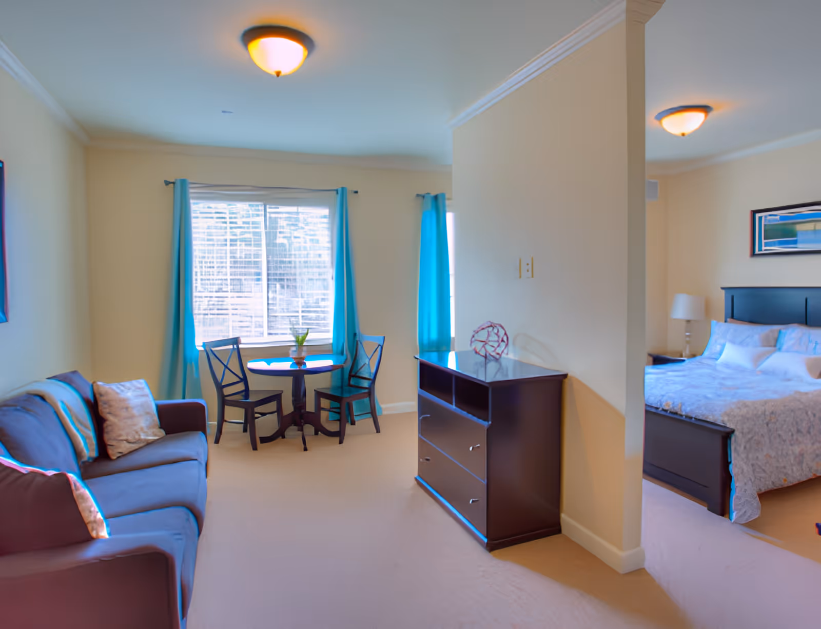 Interior view of a senior living facility room showing a living area with a dark sofa and a small round table with two chairs near a window with blue curtains. Adjacent to the living area is a bedroom with a bed, nightstand, and lamp, partially separated by a wall. The room has beige walls and carpeted floors.