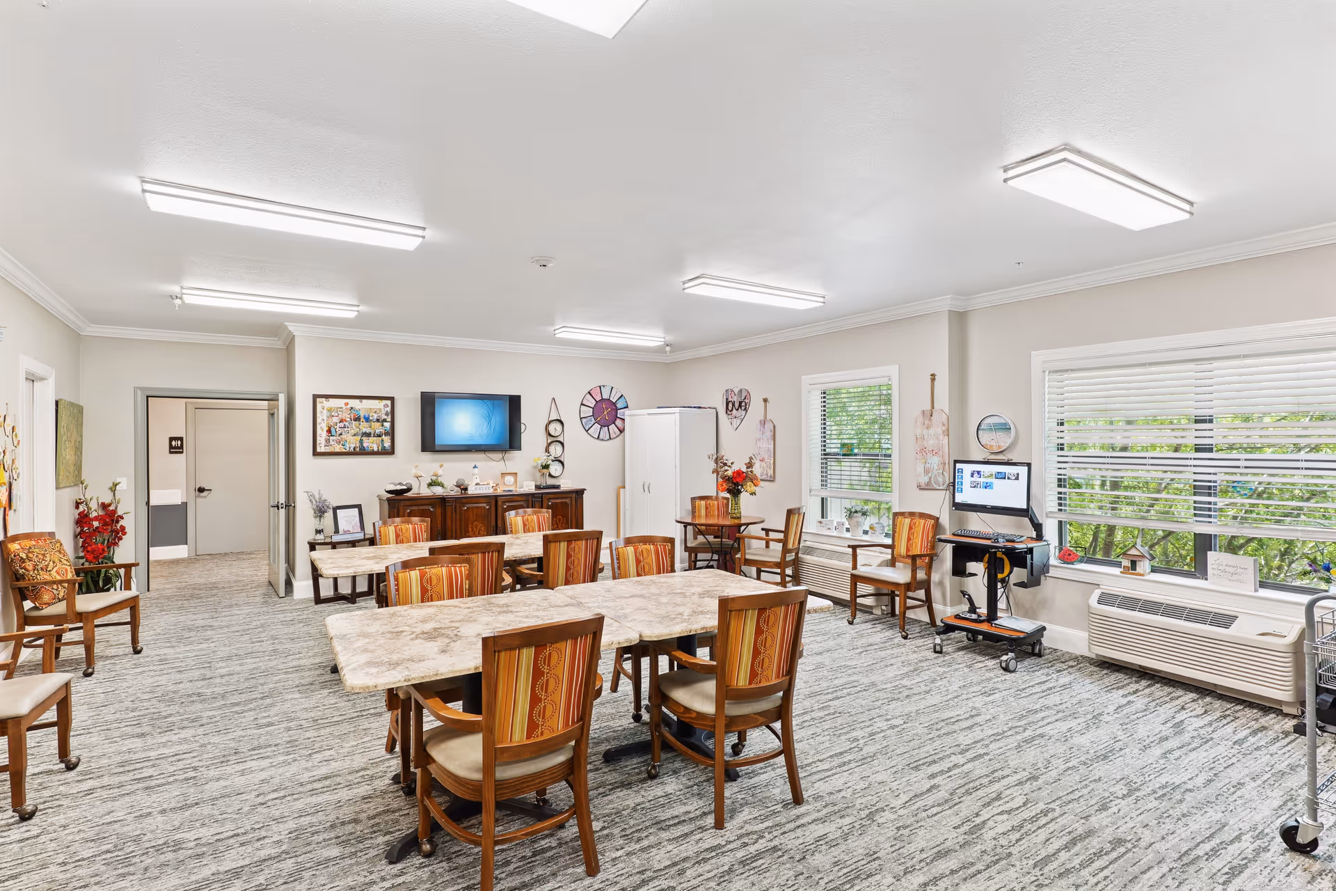 A bright, spacious common room with multiple tables and chairs arranged for group activities or dining. The room features large windows with blinds, a wall-mounted TV, decorative wall art, and a computer workstation on a rolling cart. The carpeted floor and neutral-colored walls create a welcoming atmosphere.
