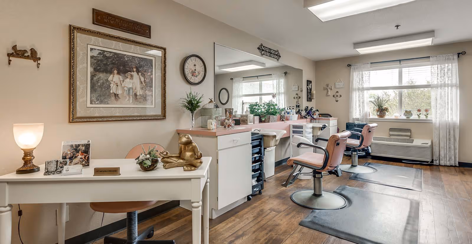 Interior view of a salon area in a senior living facility with two pink salon chairs in front of a long counter with mirrors, various plants, and hair styling tools. There is a white desk with decorative items including a lamp, a frog figurine, and framed pictures. The room has wooden flooring, a large window with sheer curtains, and soft neutral-colored walls.