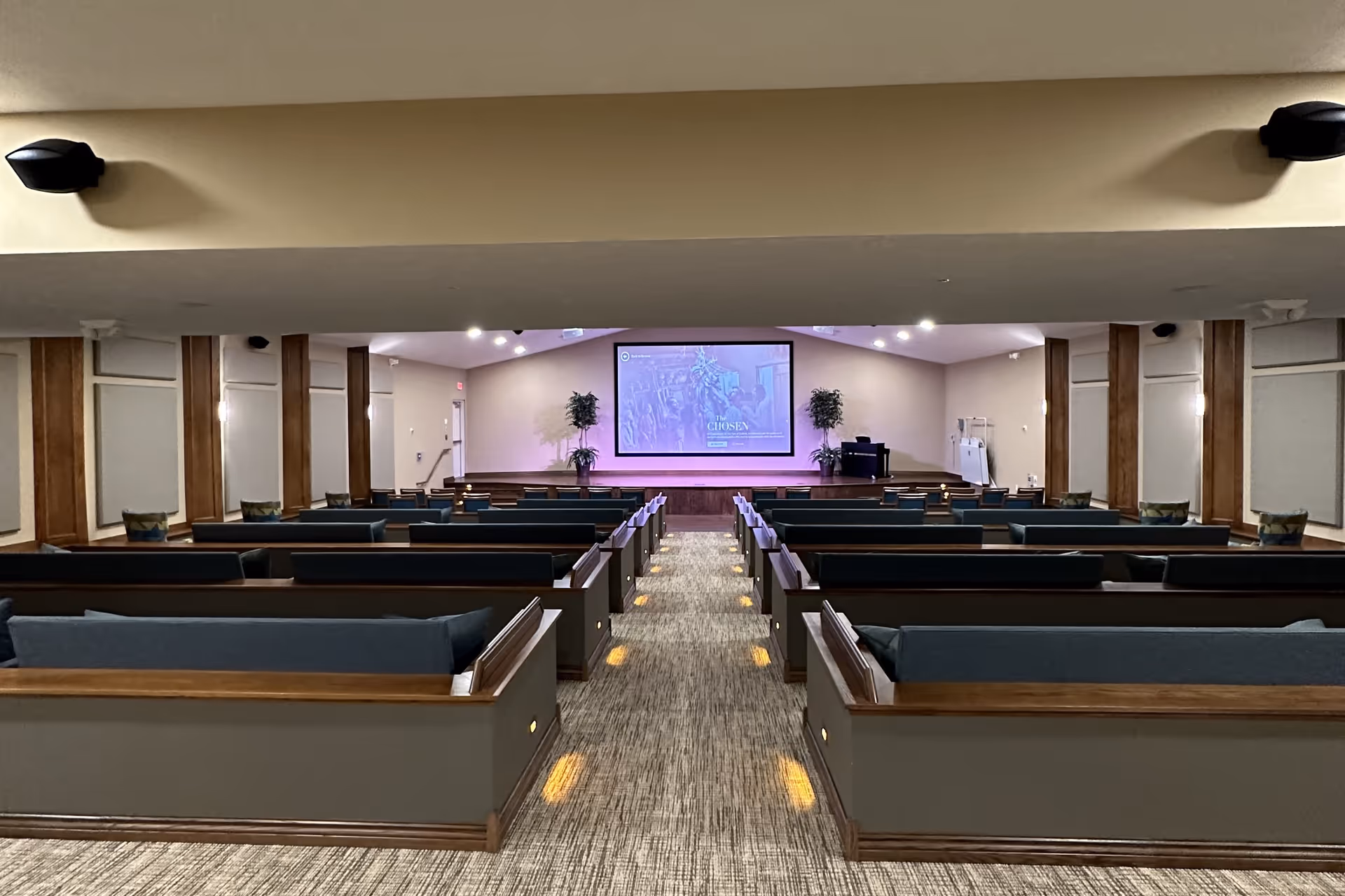 Interior view of a large room with rows of wooden pew-style seating with blue cushions, a central aisle with carpet and small lights along the sides, and a stage at the front with a large screen displaying an image and text. The room has beige walls, wooden panel accents, and two potted plants on the stage.
