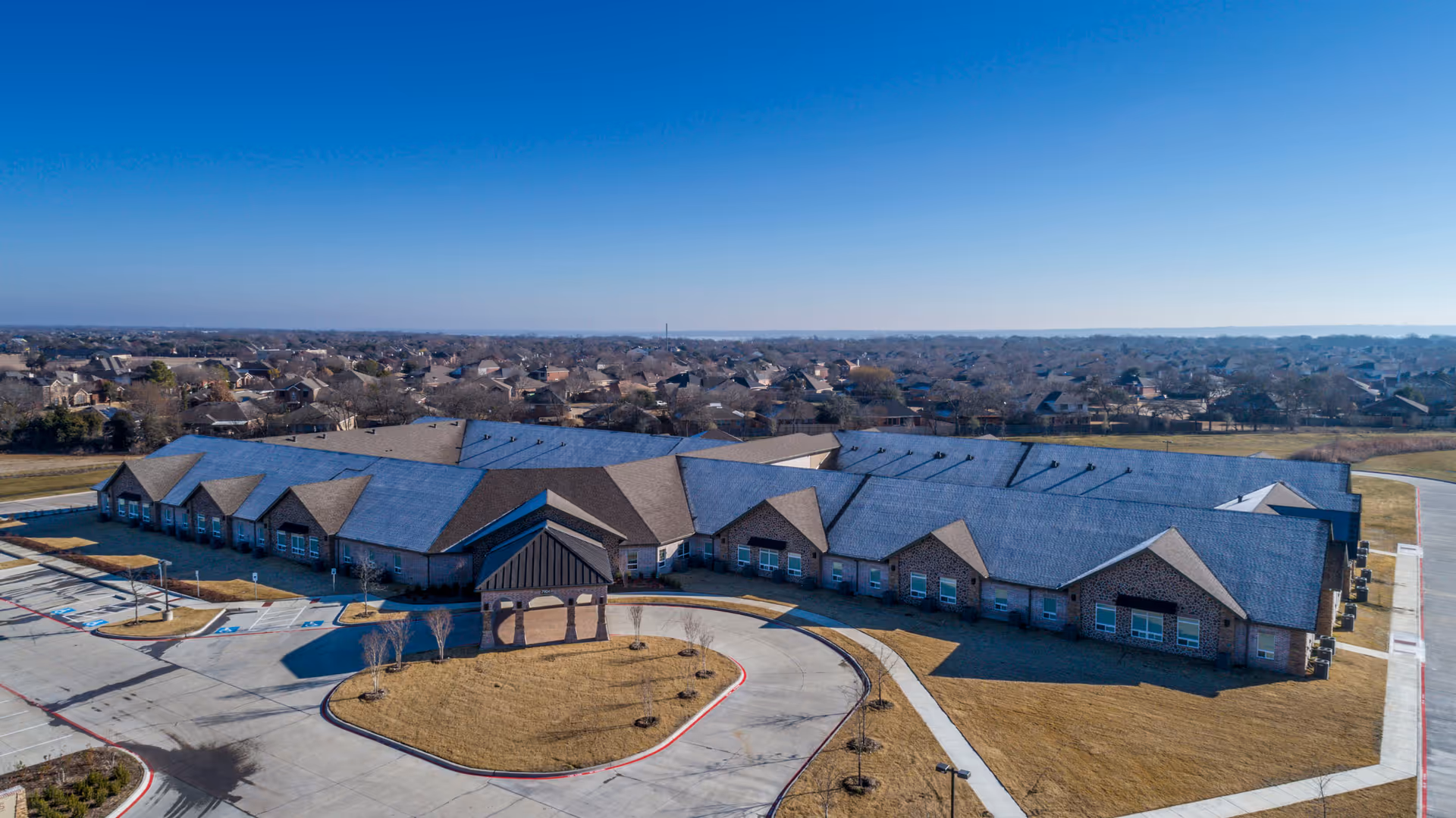 Aerial view of a single-story brick assisted living facility with a covered entrance, circular driveway, and surrounding parking in a suburban area.