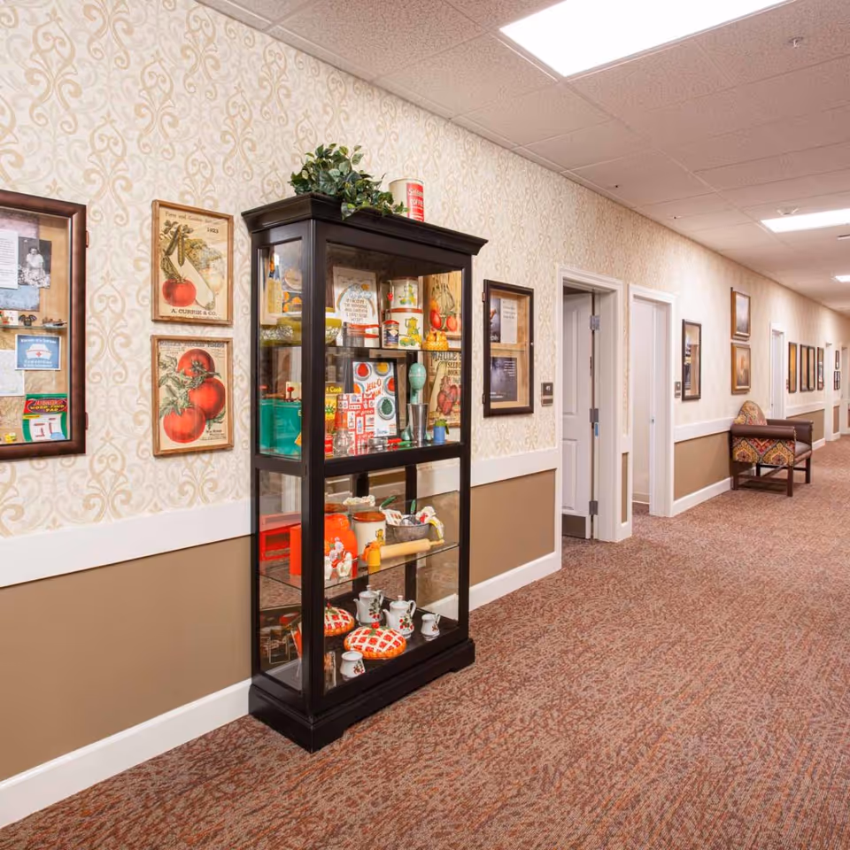 A well-lit hallway in a memory care community with patterned wallpaper and carpet. On the left side, there is a black display cabinet filled with vintage kitchen items and collectibles. The walls are decorated with framed pictures and artwork. At the far end of the hallway, there is a cushioned bench with a patterned fabric.