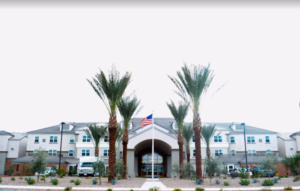 Front exterior view of Copper Springs Retirement Community building with a row of tall palm trees and an American flag in the center.